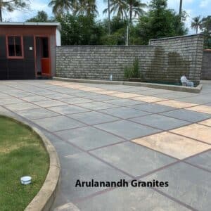Earth-toned granite entryway paved with large square gray and beige tiles in a grid pattern, featuring a curved grass strip with embedded white cylinder lights, a partially open wooden gate to a red-door building, and a stone water-feature wall with an animal-head fountain.