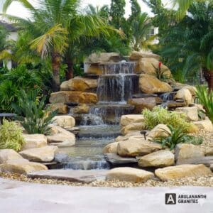 Multi-tiered granite waterfall by Arulanandh Granites, cascading over flat-rock tiers into a pond framed by gravel and tropical plants.