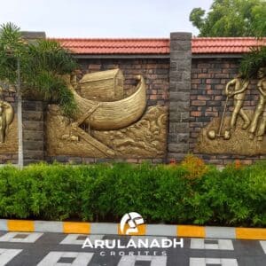 Frontal view of a sculpted granite wall with golden relief panels, dark brick columns, red-tiled roof, and landscaped foreground with palm trees and checkered paving.