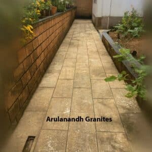 Narrow stone-paved garden pathway installed ten years ago, bordered by a brick wall with potted plants on the left and a raised garden bed on the right.